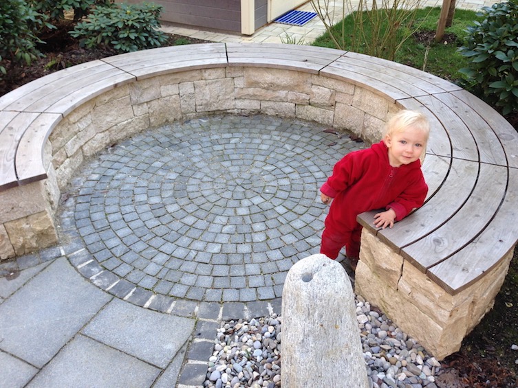 Sitzbank aus Jura Rebmauerwerk mit Sitzauflage aus L&auml;rche in runden Segmenten. Belag dunkler Granit in Kleinstein und Platten. Einfassung mit Basalt. Neben der Bank befindet sich ein Quellstein. (M&uuml;nchen)