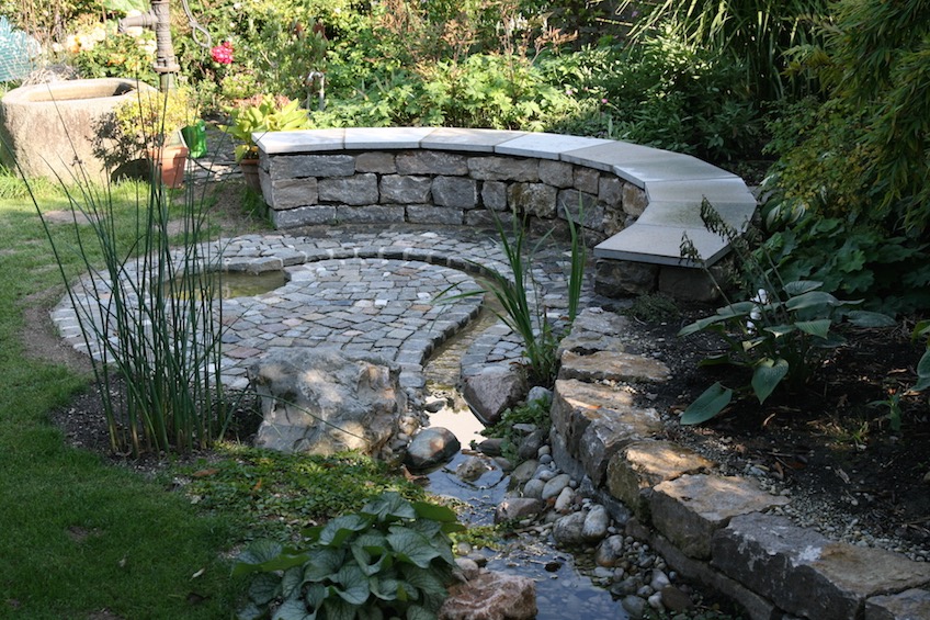 runde Bank aus Muschelkalk Mauersteinen mit Abdeckplatten aus Muschelkalk. Die Fläche wurde mit einer Steinmischung aus Granit, Porphyr, Sandstein, Marmor und Basalt, befestigt. (Moosburg)