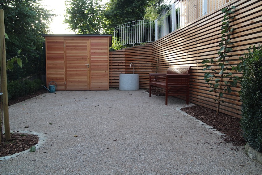 Die Fläche wurde mit einer wassergebundenen Decke, oder auch Mineralbeton genannt, befestigt. Die Betonwand wurde mit einer waagrechten Lattung aus österreichischer Gebirgslärche verkleidet. Dazu passend der Gartenschrank. Auf der Fläche befindet sich ein Granittrog mit Auslauf aus Edelstahl, sowie Obstgehölze in Spalierform, frei wachsend und Buchs. (Zolling bei Freising)