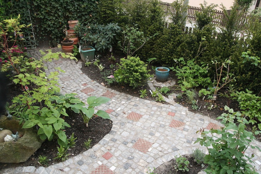 Weg aus buntem gemischten Pflaster. Gepflasterte Ranken aus Granit Kleinstein zum Stellen von Töpfen. Üppige Schattenbepflanzung. (Freising)