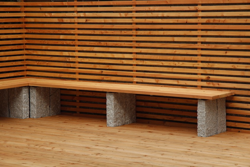 Mit dem Sichtschutz aus Lärche wurde eine Garagenwand verkleidet. Davor wurde eine Holzterrasse gelegt, die über eine Teichfläche ragt. Eine Sitzbank mit Holzauflage lädt zum Verweilen ein. (München)