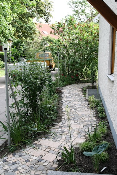 Geschwungener Weg aus buntem Pflaster. Zaun aus Metallpfosten mit Edelstahlseilen, die mit Kletterpflanzen berankt wurden. Schattenpflanzung. (Garching bei München)