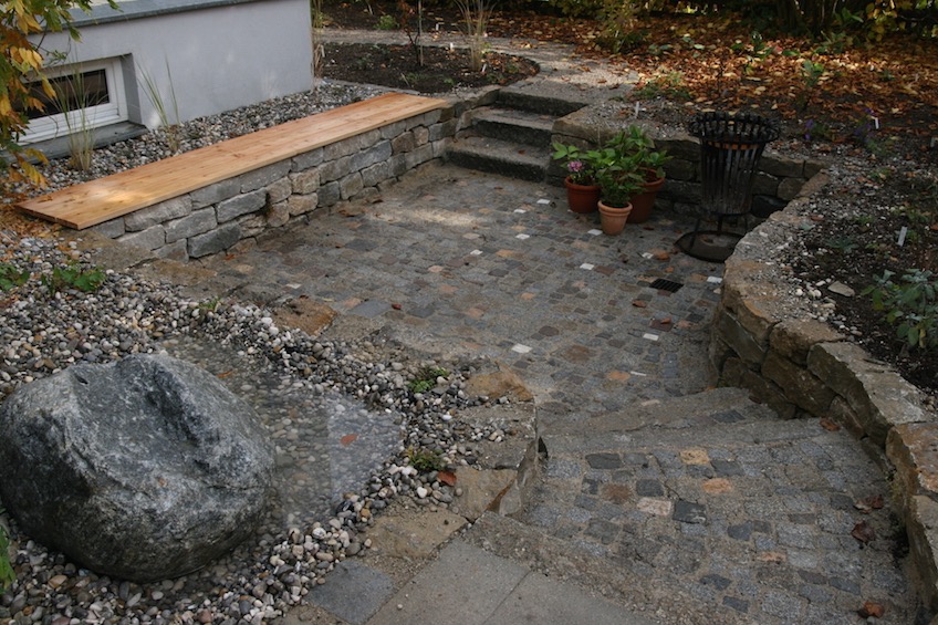 Kleiner Sitzplatz mit Bank und Quersten, der 50cm tiefer als das ihn umgebende Niveau liegt. Die Mauern sind aus Muschelkalk, der Belag ist gemischt. (München)