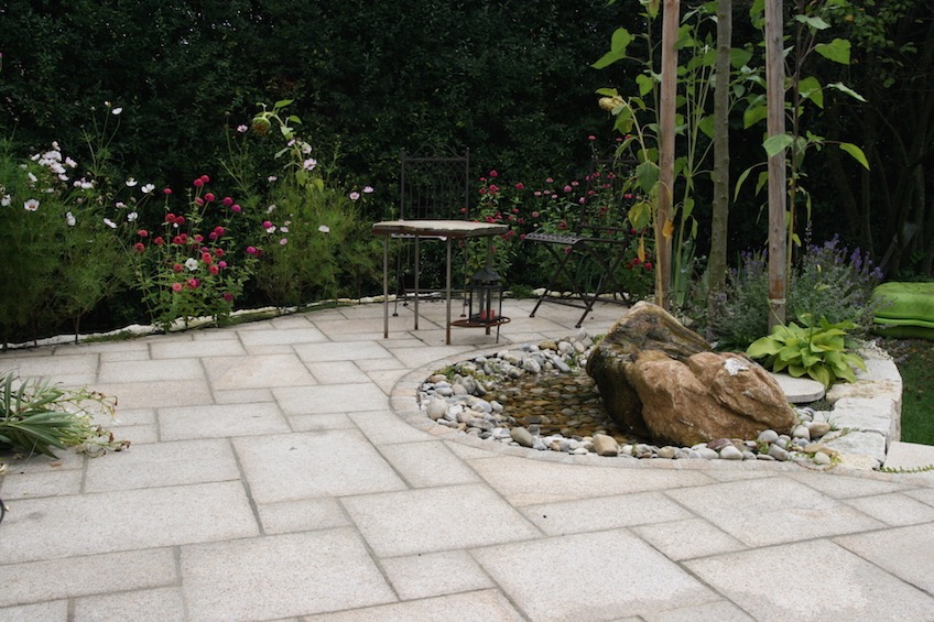 Terrasse mit organischen Schwüngen und Quellstein mit sichtbarer Wasserfläche. (Pulling bei Freising)